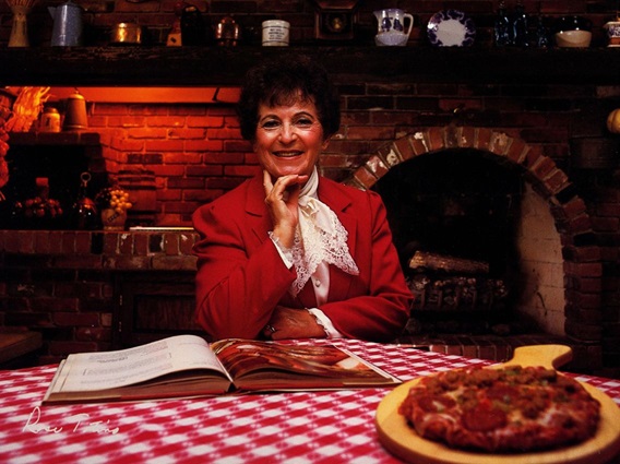 Rose Totino with her hand on her chin smiling, sitting at a table with a red and white checkered tablecloth with an open recipe book and cooked pizza sitting in front of her. In the background you can see the dimly lit brick pizza oven.