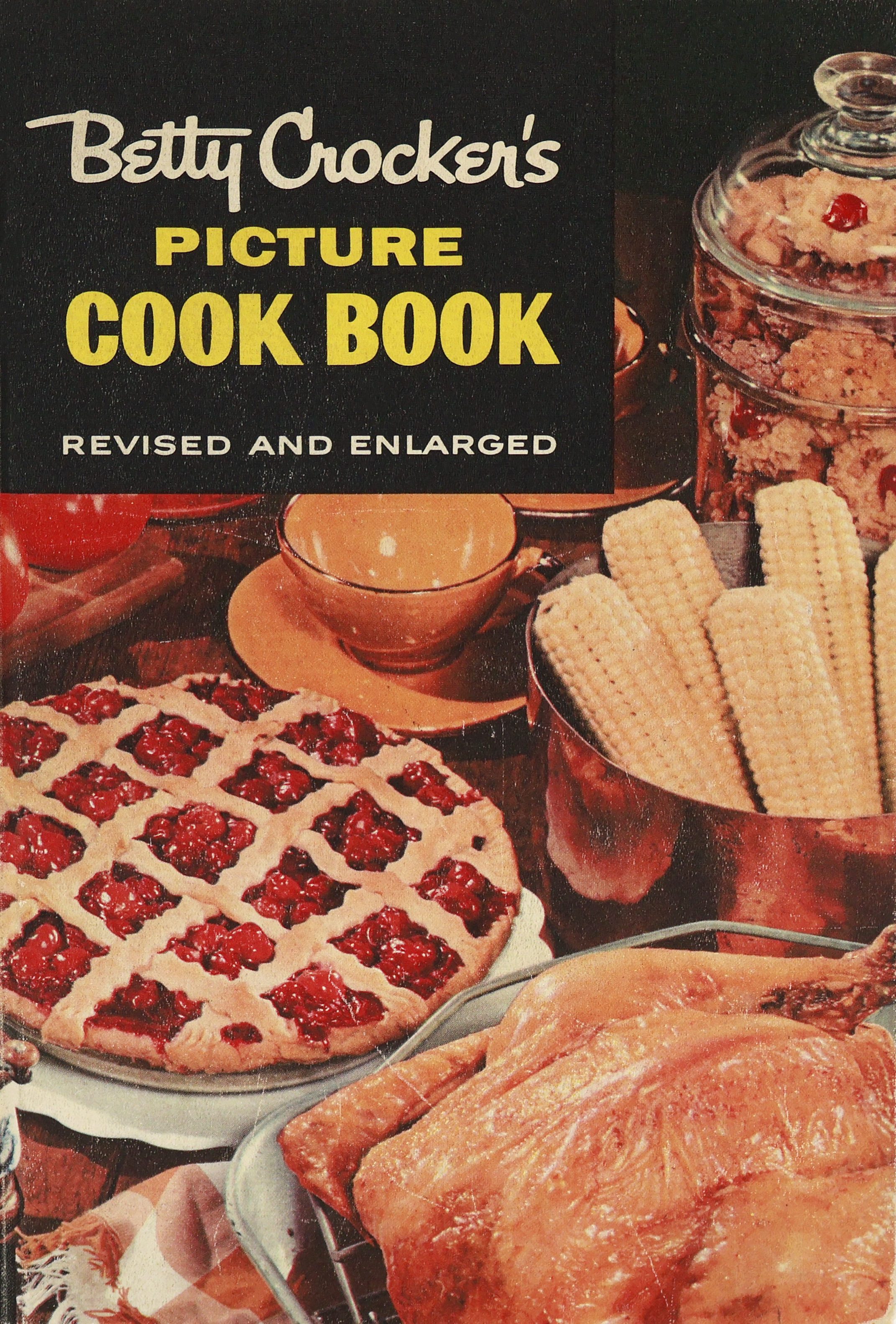 o	Cover of the 1956 Betty Crocker Cookbook with a black upper portion with “Picture Cookbook” written in yellow. Front has a cherry pie, whole turkey, copper kettle with corn on the cob and a glass cookie jar with cookies. 