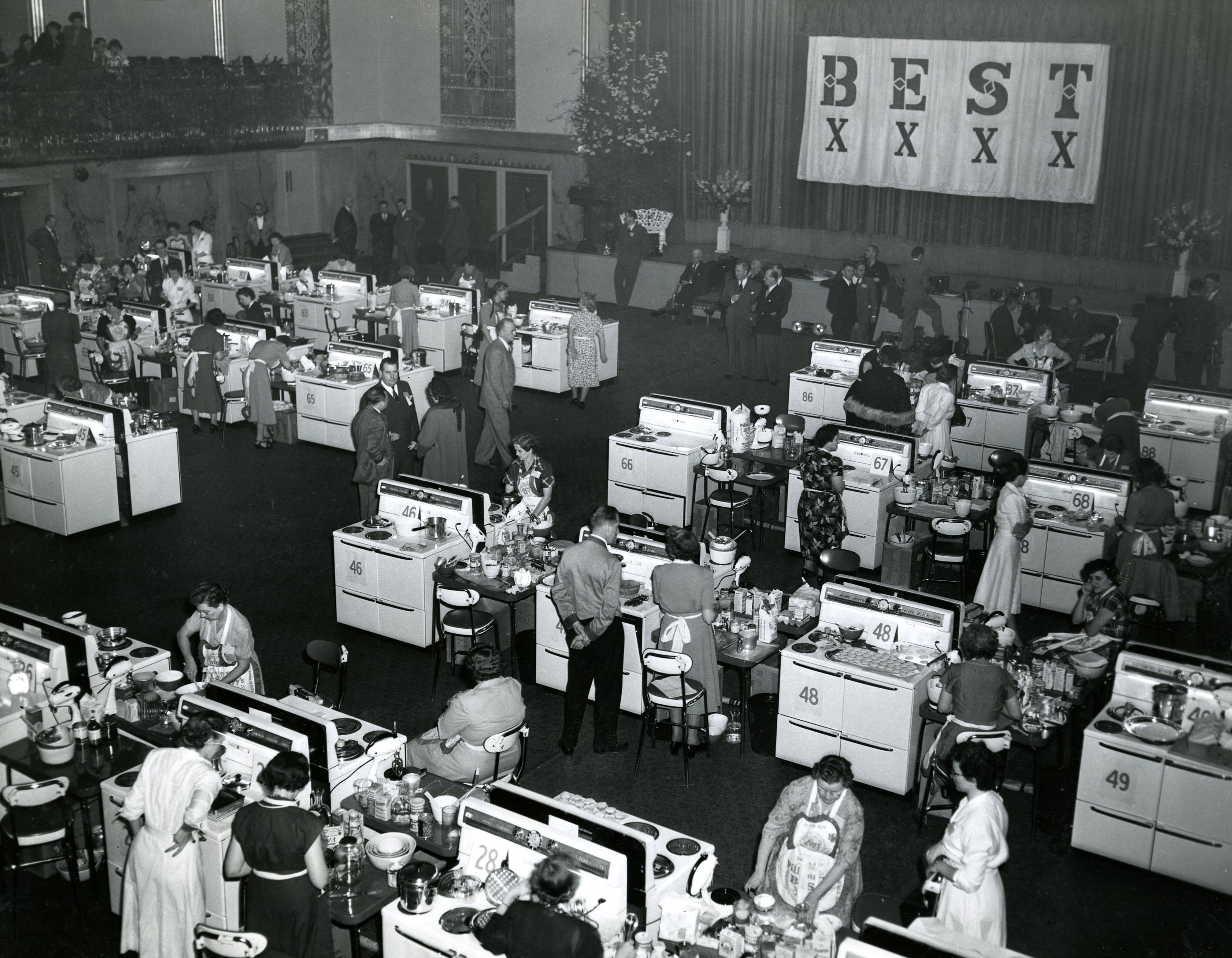 A historic, black-and-white, high-angle photograph of the Pillsbury Bake-Off contest floor. The large hall is filled with dozens of contestants, primarily women, standing at their individual baking stations, each equipped with a white stove and prep table. A large banner hangs in the background that reads "BEST".