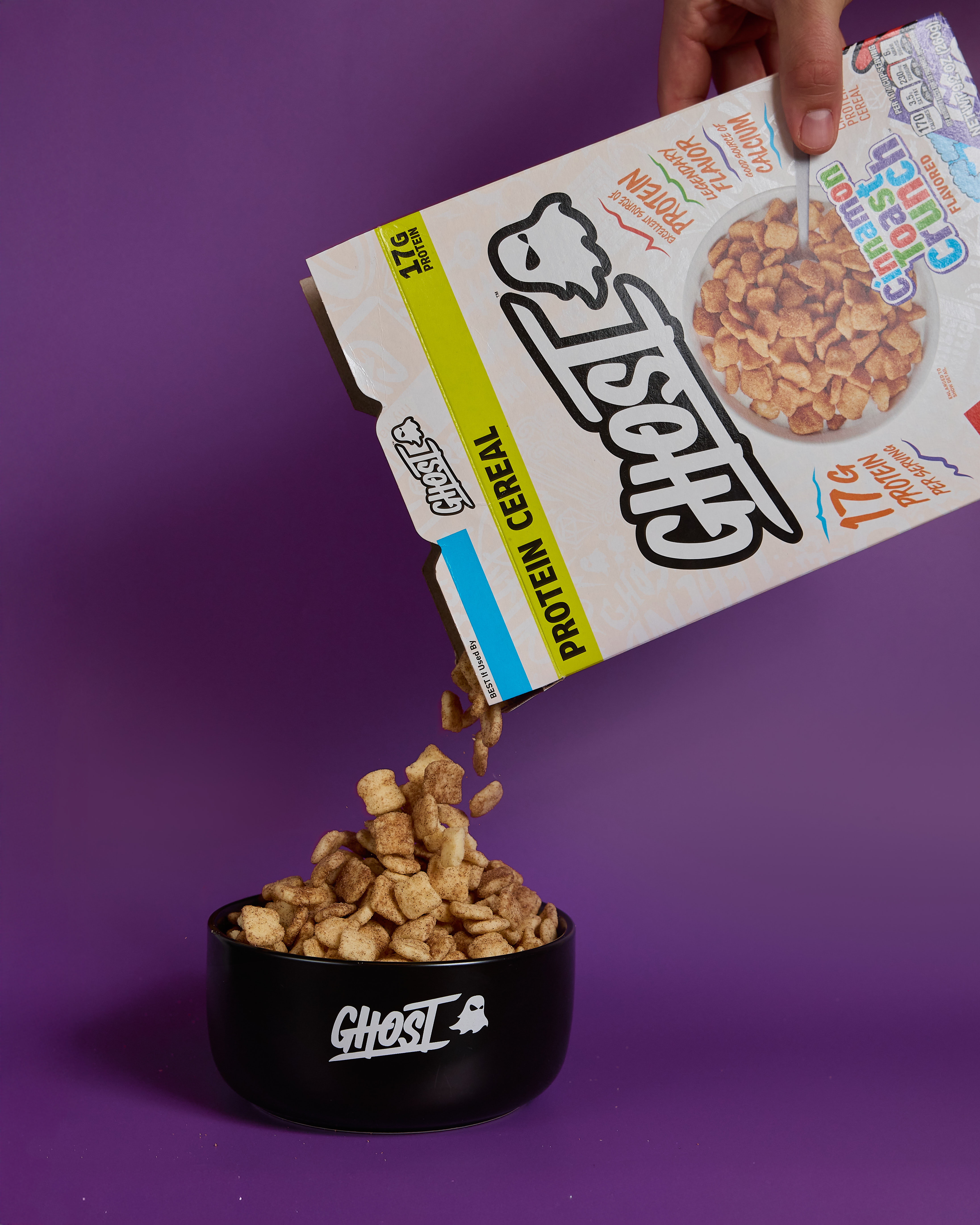 GHOST® PROTEIN CEREAL x CINNAMON TOAST CRUNCH™ being poured into a black bowl with a purple background.